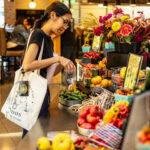 A young woman wearing glasses and a tote bag looks over a farm market table.