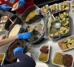 People wearing blue gloves work to fill to-go containers with food.