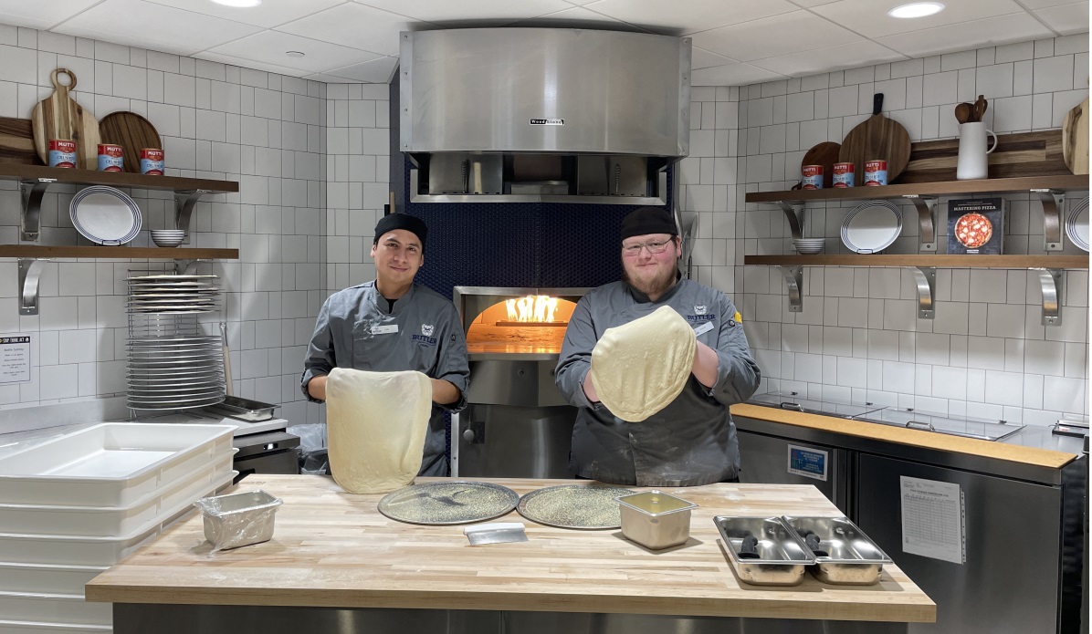 Two men holding pizza dough stand in front of a commercial pizza oven