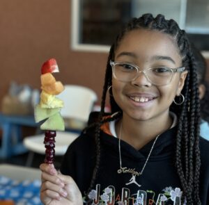 A smiling young girl with braids in her hair holds up a fruit kebab with melon, grapes, and strawberries.