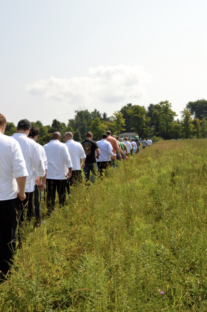 A group of people wearing white chefs' coats walk single file through a field.